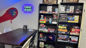Una habitación con una mesa de madera, un letrero de neón que dice 'GAME ROOM' y estantes llenos de juegos de mesa.