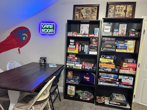 A room with a wooden table, a neon sign reading 'GAME ROOM', and shelves filled with board games.