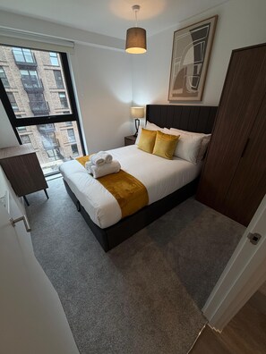 Ein modernes Hotelzimmer mit einem großen Bett, einem Nachttisch, einer Lampe und Blick auf die Stadt durch ein großes Fenster.