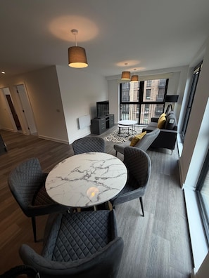 Un salon moderne avec une table ronde en marbre, des chaises noires rembourrées, un canap é avec des coussins et une grande fenêtre donnant sur des immeubles.