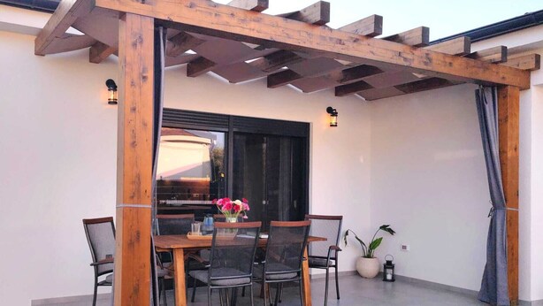 En udendørs terrasse med et træbeklædt pergolaoverdækning, et bord med stole og en potteplante.