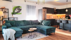 En moderne stue med en hjørnesofa, et lille rundt bord og et køkken i baggrunden.