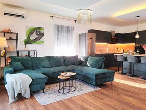 Ein modernes Wohnzimmer mit einem Ecksofa, einem kleinen runden Tisch und einer Küchenzeile im Hintergrund.
