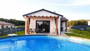 En swimmingpool med beboelse, overdækket terrasse, indhegnet have og et hus i baggrunden.