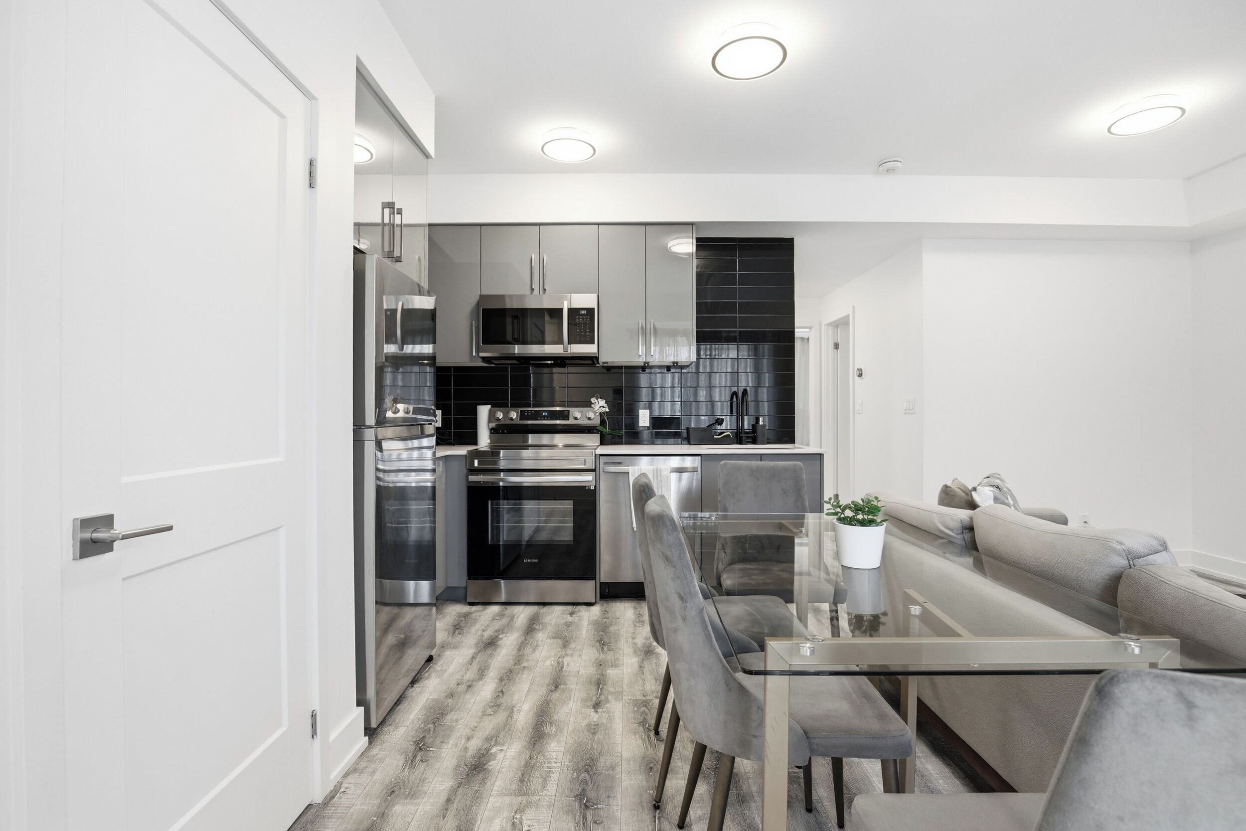 A modern kitchen with stainless steel appliances, a dining area with a glass table, and a grey sofa.