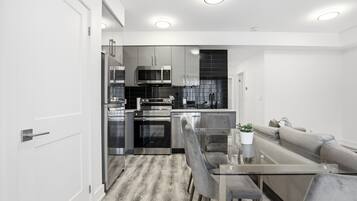 A modern kitchen with stainless steel appliances, a dining area with a glass table, and a grey sofa.