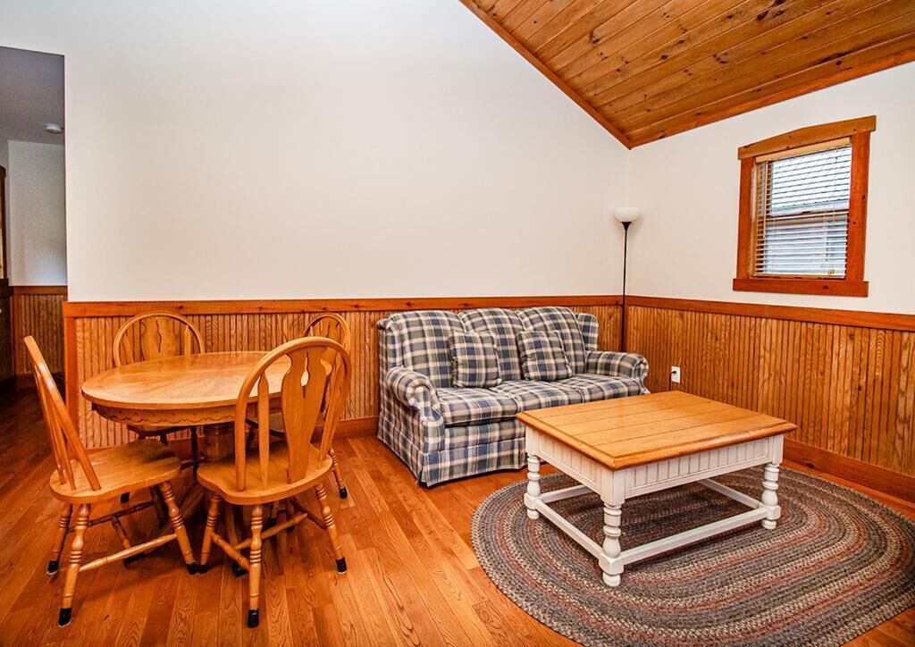 A room with a wooden dining table and chairs, a plaid sofa, and a small window with blinds.