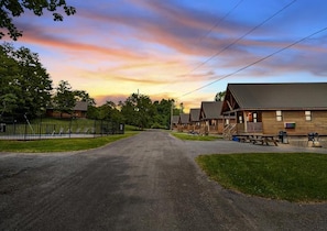 A campground with multiple cabins, a paved driveway, picnic tables, and a fenced playground area. - Chalet Village (Milford)