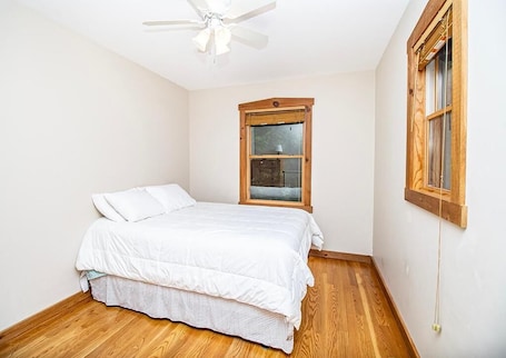 A bedroom with a bed, wooden flooring, a ceiling fan, and a window with a wooden frame.
