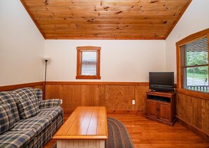 Una habitación con techo de madera, un televisor sobre un mueble de madera, un sofá escocés y una ventana con persianas.
