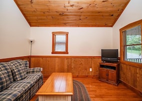 A room with a wooden ceiling, a television on a wooden stand, a plaid sofa, and a window with blinds.