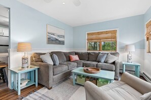 A living room with a grey sofa, two side tables, a coffee table, and a window with blinds. - Pirate's Cove Cottage | beach, mini golf, backyard (Old Orchard Beach)