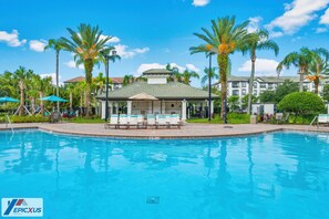 A swimming pool with lounge chairs and umbrellas, surrounded by palm trees and a building with a green roof. - The Epic 36208 Lake View Vista Cay Resort Orlando Florida (Orlando)