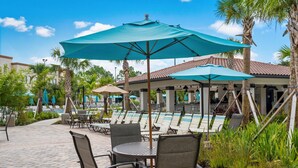 A poolside area with lounge chairs and umbrellas, surrounded by palm trees and a building with a tiled roof. - The Epic 48108 Lake View Vista Cay Resort Orlando Florida (Orlando)
