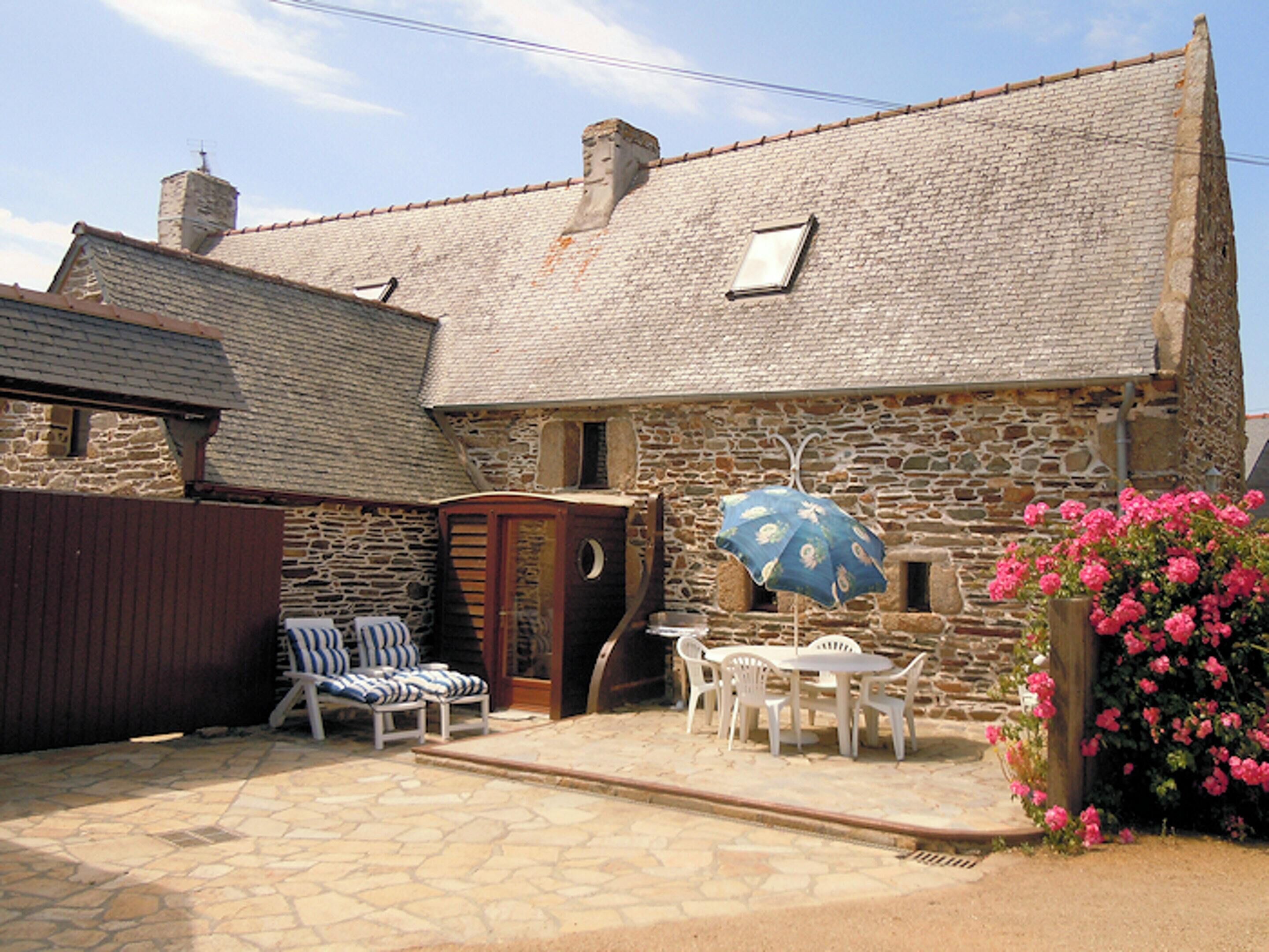 Une maison en pierre avec un toit en tuiles, une petite fenêtre et une porte. On y trouve une terrasse avec une table et des chaises, ainsi qu’un parasol bleu.
