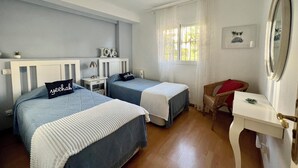 Ein Schlafzimmer mit zwei Betten, einem Deckenventilator, einem Fenster mit Vorhängen, einem kleinen Tisch und einem Stuhl.