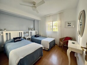 Une chambre avec deux lits, un ventilateur de plafond, une fenêtre avec des rideaux, une petite table et une chaise.