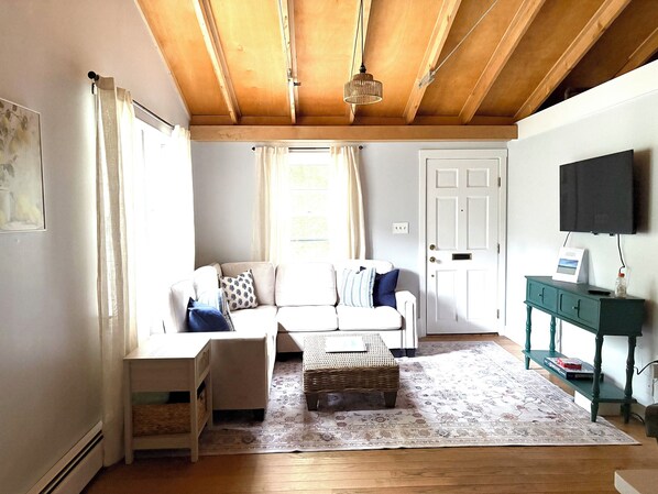Un salon avec un plafond en bois, un canapé blanc, une petite table, un pouf en osier et une console verte sur laquelle se trouve un téléviseur.
