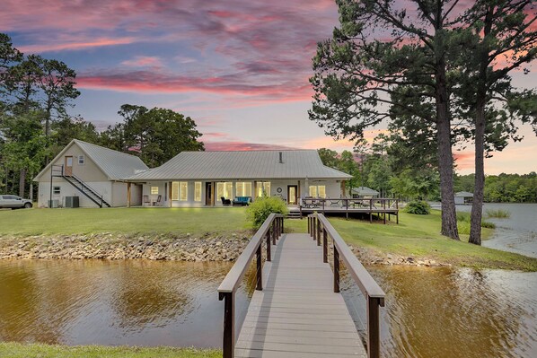 A two-story building with a dock leading to a body of water, surrounded by trees and a grassy area. - Two homes- 5 bedrooms on beautiful private lake. (Long Branch)