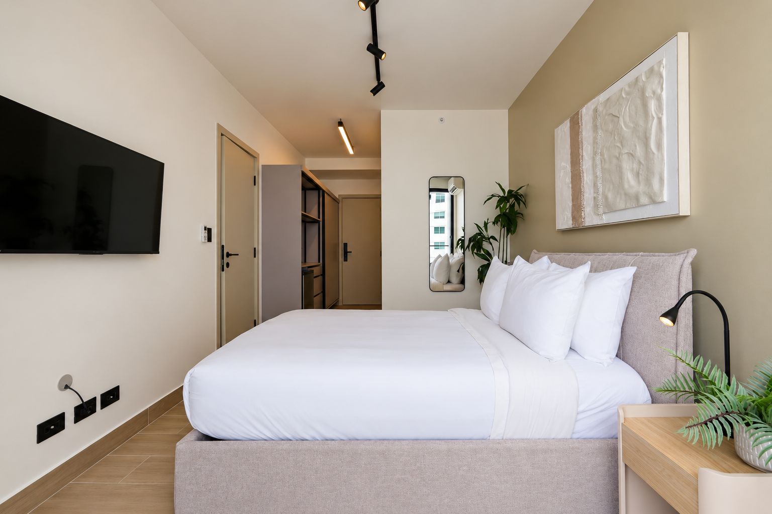 A modern bedroom with a large bed, a flat-screen TV mounted on the wall, a bedside table with a lamp, and a mirror.