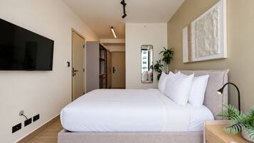A modern bedroom with a large bed, a flat-screen TV mounted on the wall, a bedside table with a lamp, and a mirror.