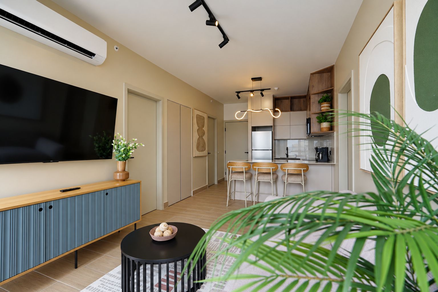 A modern living room with a flat-screen TV, a blue cabinet, a round coffee table, and a kitchen area with bar stools.