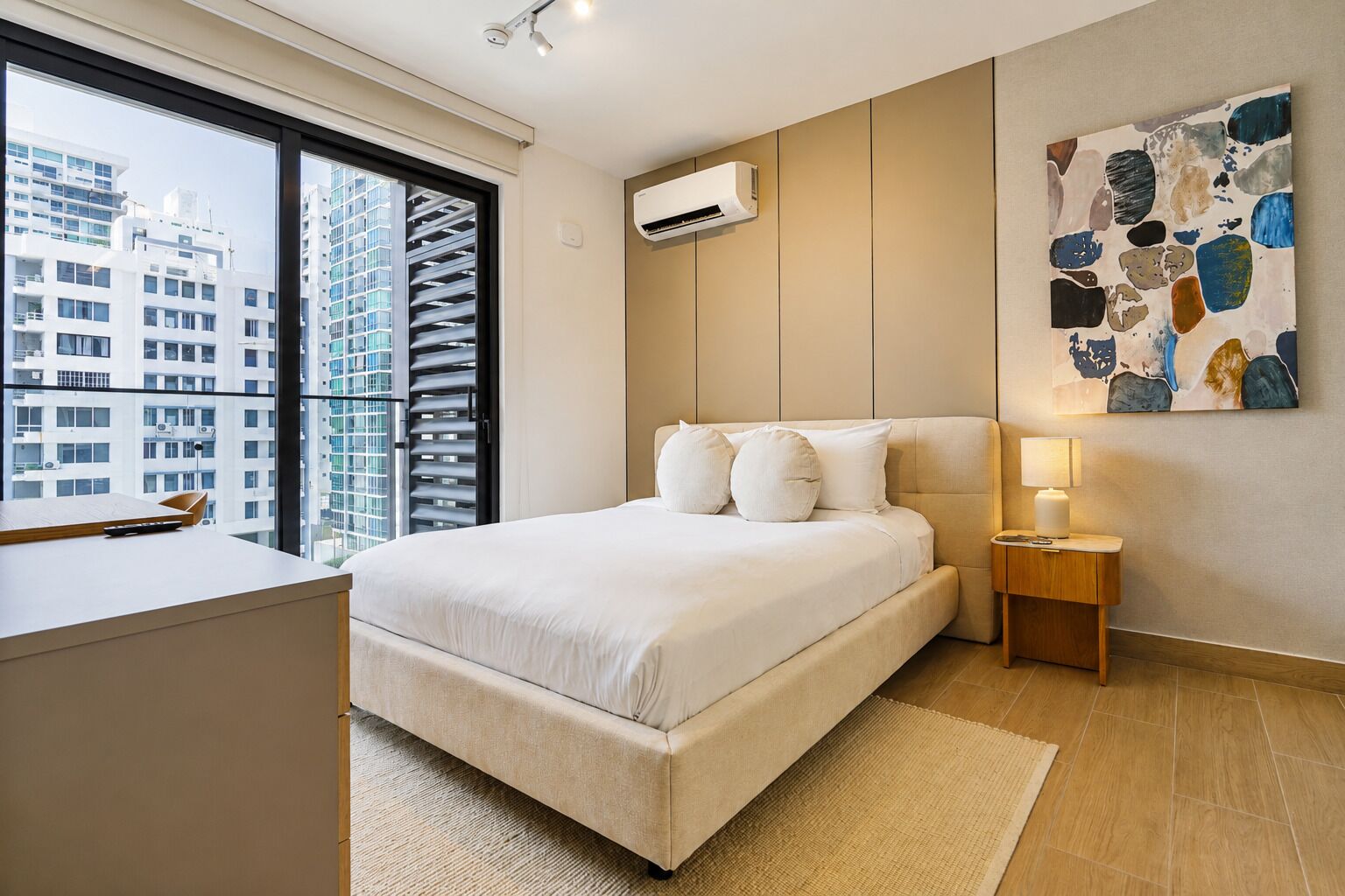 A modern bedroom with a large bed, a desk, and a view of high-rise buildings.