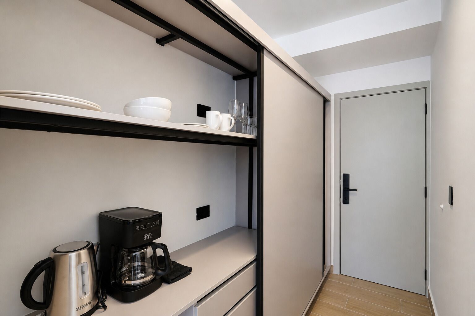 A modern kitchen with a built-in coffee maker and a kettle, a white door, and a shelf with dishes and glasses.