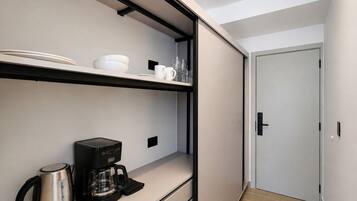 A modern kitchen with a built-in coffee maker and a kettle, a white door, and a shelf with dishes and glasses.