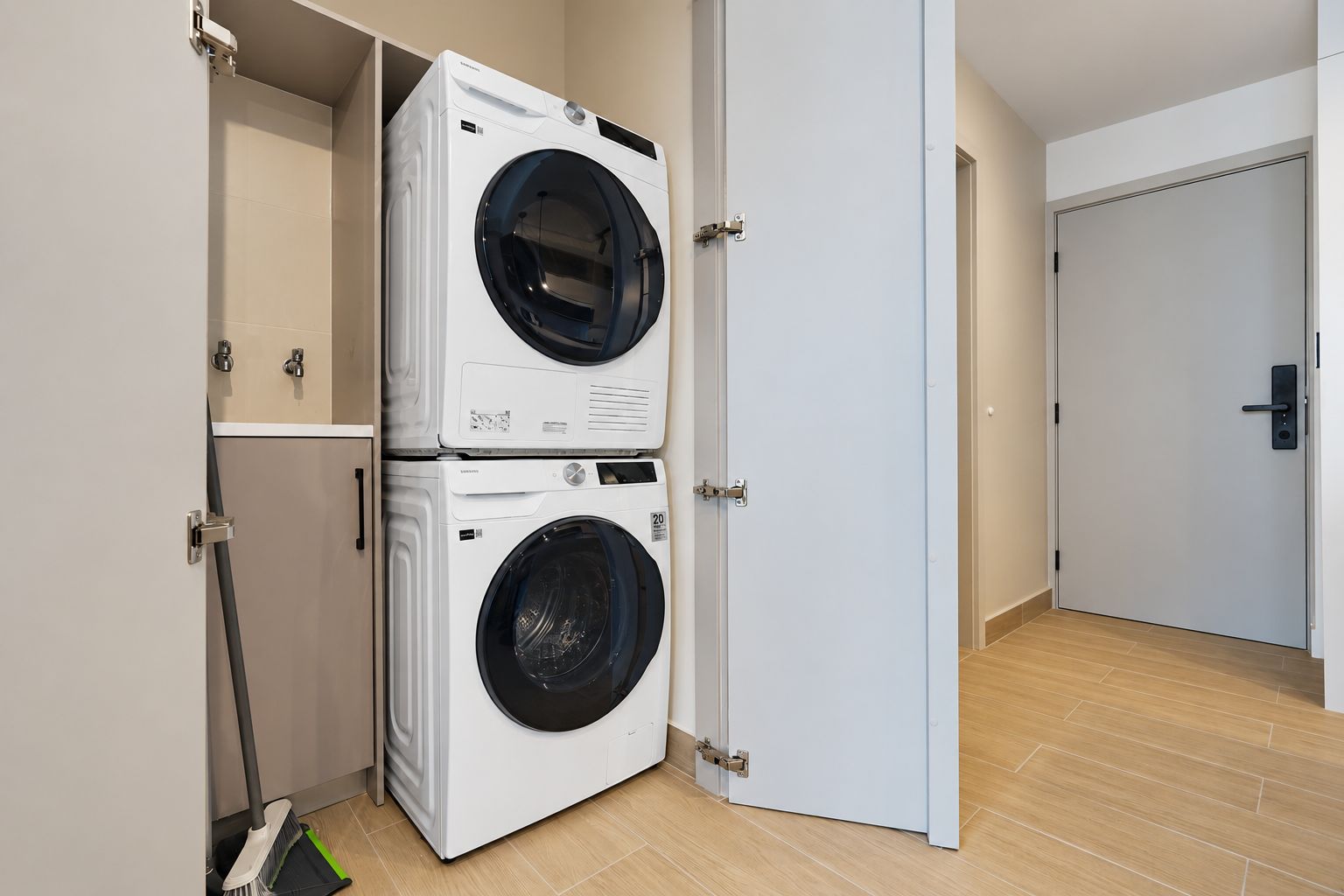 Un cuarto de lavandería con dos lavadoras de carga frontal apiladas, un escoba y una puerta que da a otra habitación.