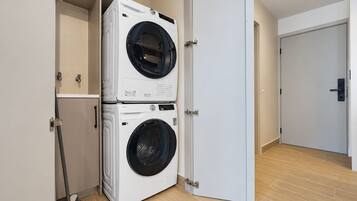 Un cuarto de lavandería con dos lavadoras de carga frontal apiladas, un escoba y una puerta que da a otra habitación.