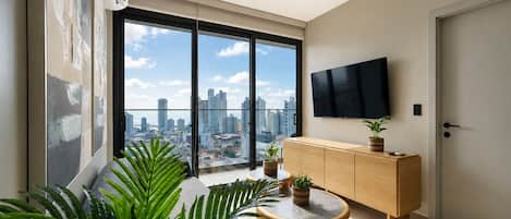 A modern living room with a large window, a flat-screen TV, a wooden cabinet, and a round table.