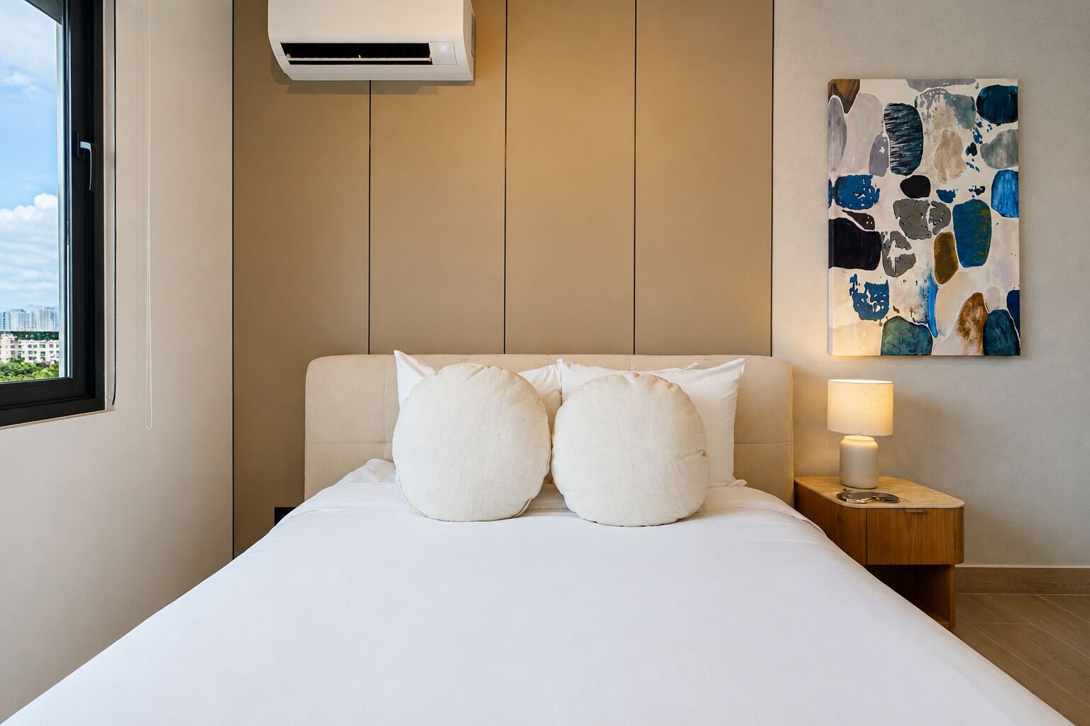 Una habitación de hotel con una cama, dos almohadas, una mesita de noche con una lámpara, un equipo de aire acondicionado y un cuadro en la pared.