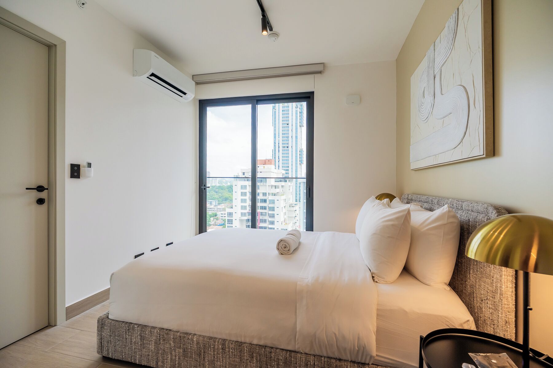 A hotel room with a bed, a window with a view of buildings, an air conditioning unit, and a wall-mounted artwork.