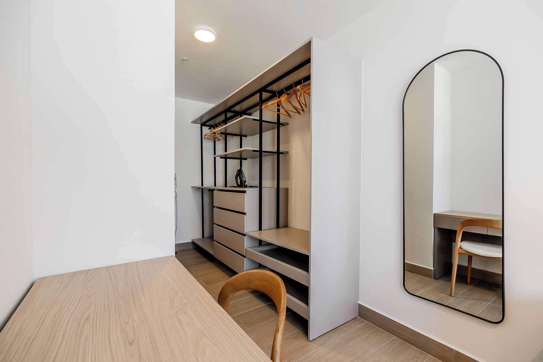 A compact room with a wooden table, a chair, a wardrobe with shelves, and a mirror on the wall.
