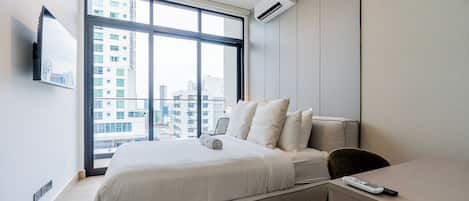 A modern hotel room with a large bed, a desk, and a view of the city through floor-to-ceiling windows.