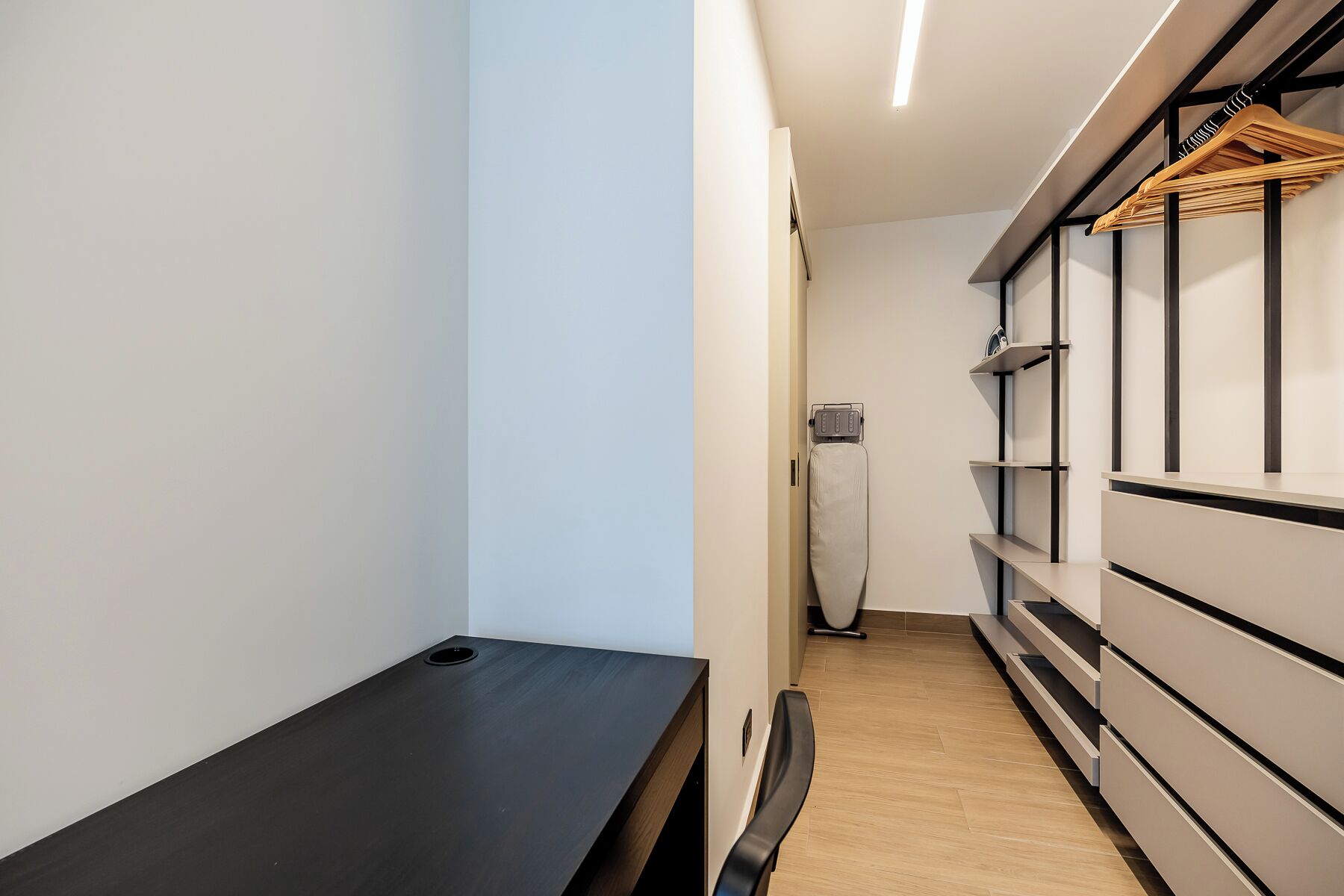 Un couloir étroit, avec un banc noir sur la gauche, un mur blanc et un porte-manteau en bois avec des cintres sur la droite.