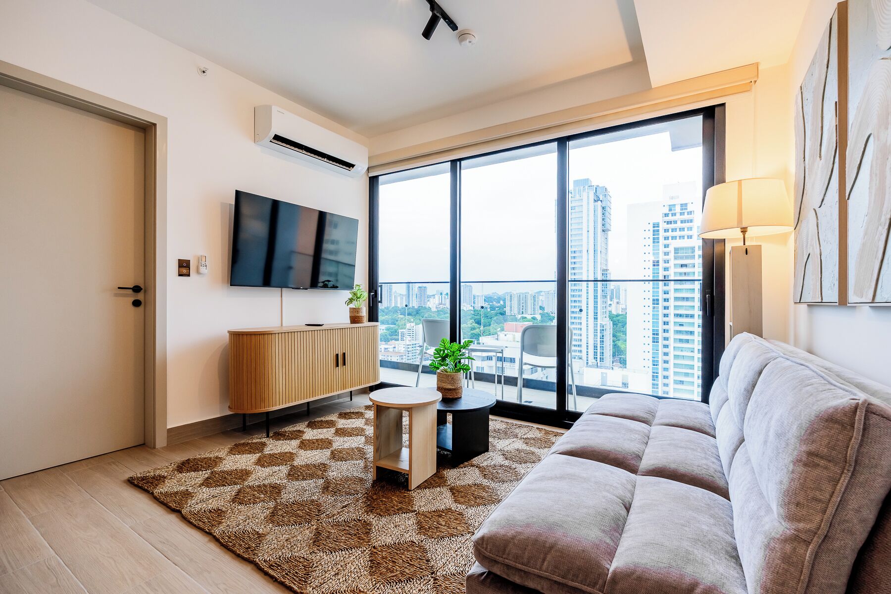 Uma sala de estar moderna com uma televisão de tela plana, um sofá, uma mesinha redonda e uma grande janela com vista para a cidade.
