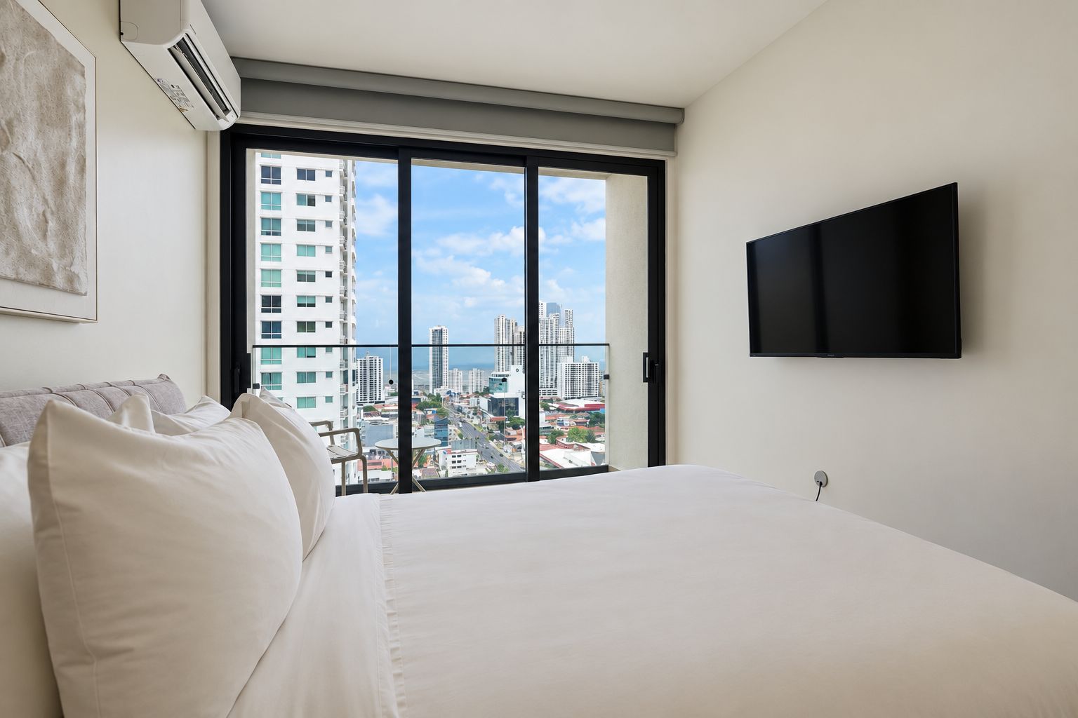 Ein Hotelzimmer mit Bett, Flachbildfernseher und Blick über die Stadt durch ein großes Fenster.