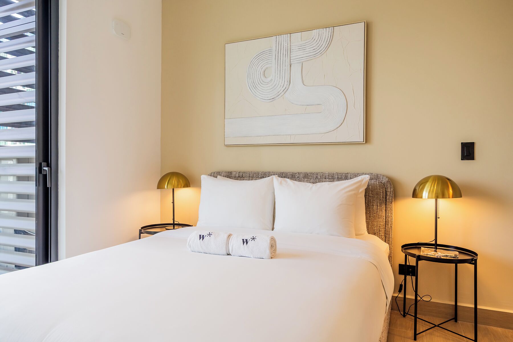 Habitación de hotel con cama, dos lámparas de noche, una obra de arte en la pared y una ventana con persianas.