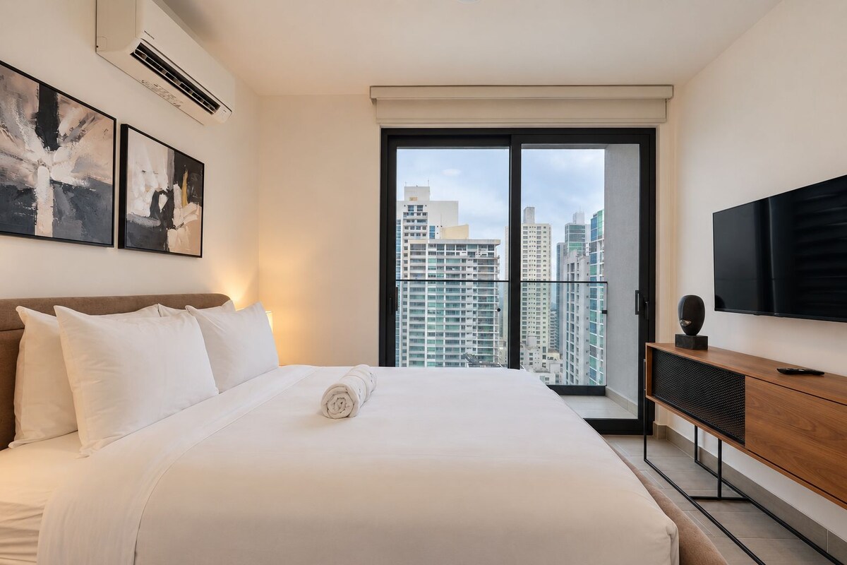 A hotel room with a large bed, a flat-screen TV, a wooden dresser, and a city view through a large window.