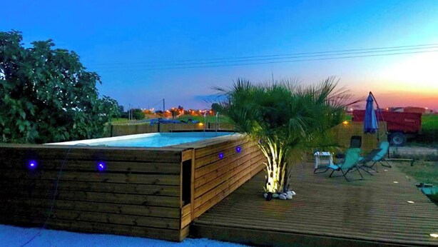 Une terrasse en bois avec une piscine, entourée de verdure, et un coucher de soleil en arrière-plan.