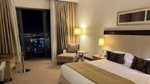 Habitación de hotel con una cama grande, una silla, una mesita y vistas a la ciudad de noche.
