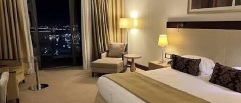 Une chambre d’hôtel avec un grand lit, une chaise, une petite table et une vue sur la ville la nuit.