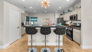 A modern kitchen with a central island, bar stools, stainless steel appliances, and decorative items on the shelves. - Entire Home + Private Casita | Private Pool & Spa | Putting Green | Fully Fenced Yard| Community Amenities (Palm Springs)