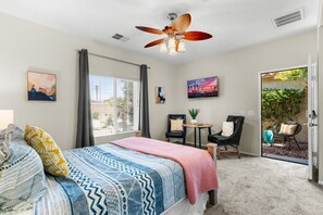 Une chambre à coucher avec un lit, un ventilateur de plafond, une fenêtre avec des rideaux, une petite table avec des chaises et une vue sur l’extérieur.