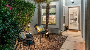 An outdoor seating area with two chairs, a palm tree, and a pathway leading to a building entrance. - Entire Home + Private Casita | Private Pool & Spa | Putting Green | Fully Fenced Yard| Community Amenities (Palm Springs)