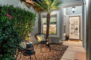 An outdoor seating area with two chairs, a palm tree, and a pathway leading to a building entrance. - Entire Home + Private Casita | Private Pool & Spa | Putting Green | Fully Fenced Yard| Community Amenities (Palm Springs)