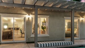 A modern outdoor pool area with a covered seating area, a glass door entrance to a living space, and a clear view of the pool. - Entire Home + Private Casita | Private Pool & Spa | Putting Green | Fully Fenced Yard| Community Amenities (Palm Springs)