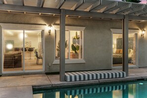 A modern outdoor pool area with a covered seating area, a glass door entrance to a living space, and a clear view of the pool. - Entire Home + Private Casita | Private Pool & Spa | Putting Green | Fully Fenced Yard| Community Amenities (Palm Springs)